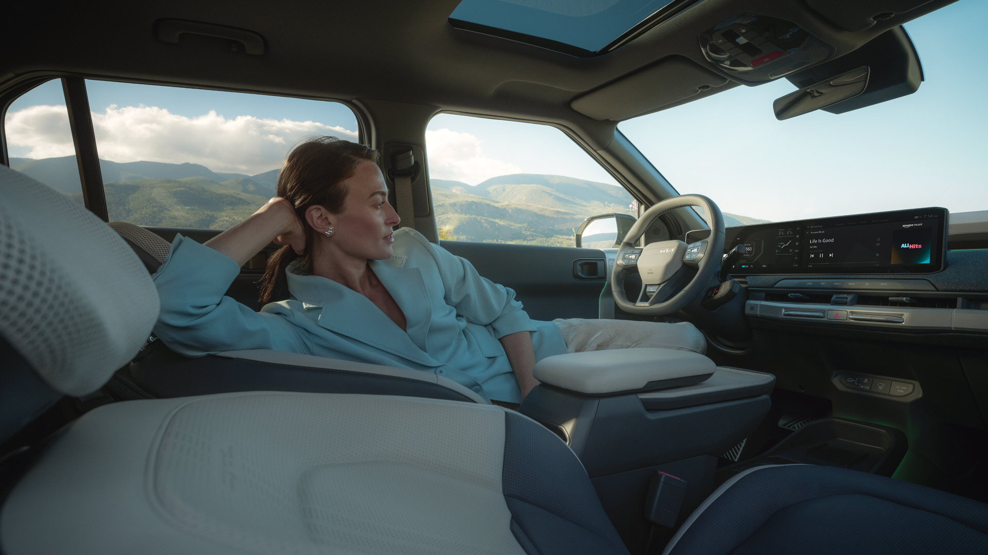 Kia EV3 - Premium Interieur mit Lounge-Charakter - Maximaler Komfort Entspannte Person im Kia EV3 Interieur mit hellem Polster und Panoramafenster vor Bergkulisse zeigt Komfort und Raumangebot