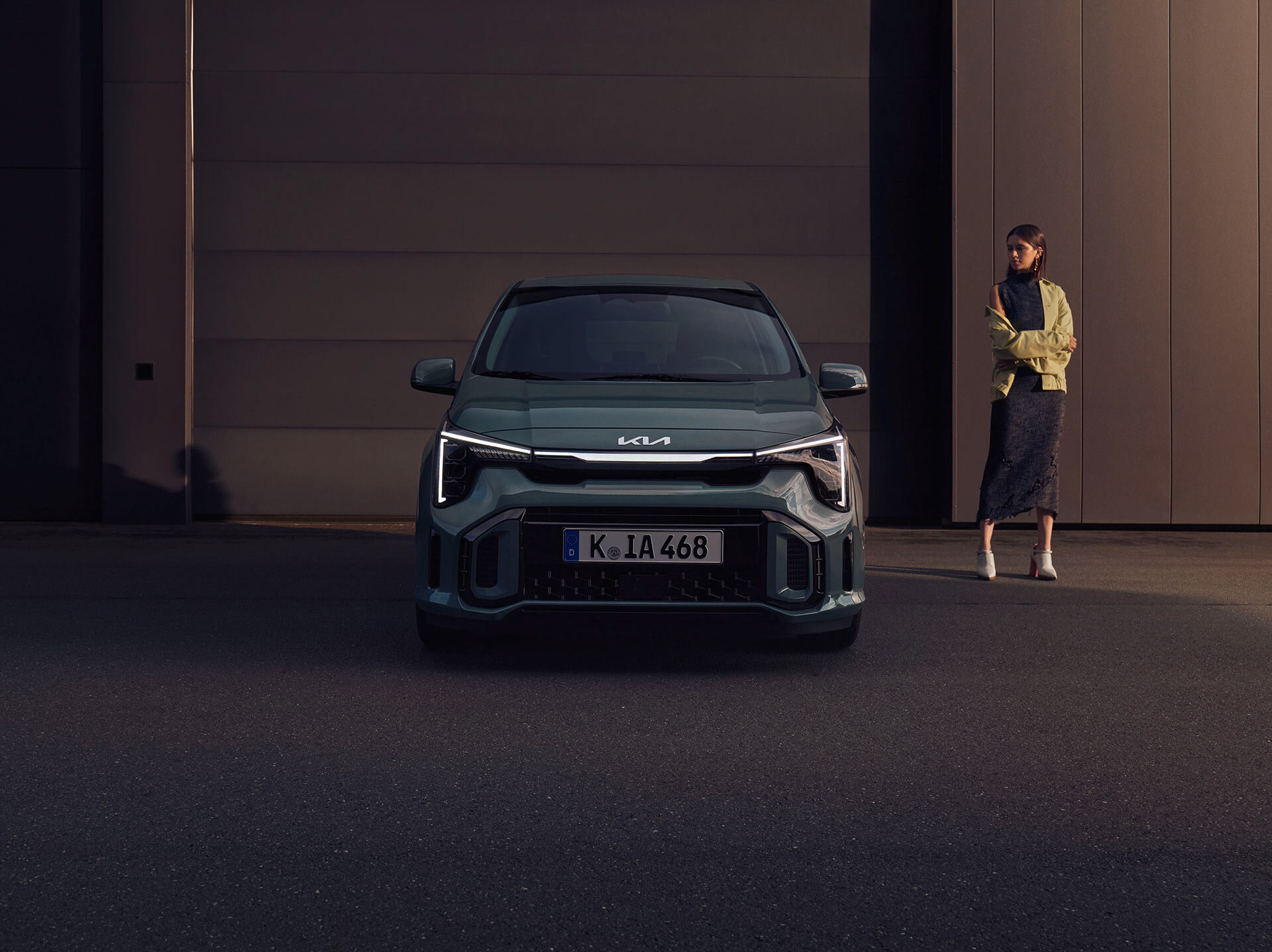 Frontansicht des Kia Picanto in der Dämmerung, markante LED-Scheinwerfer und Kia-Tiger-Face-Kühlergrill, moderne Architektur im Hintergrund