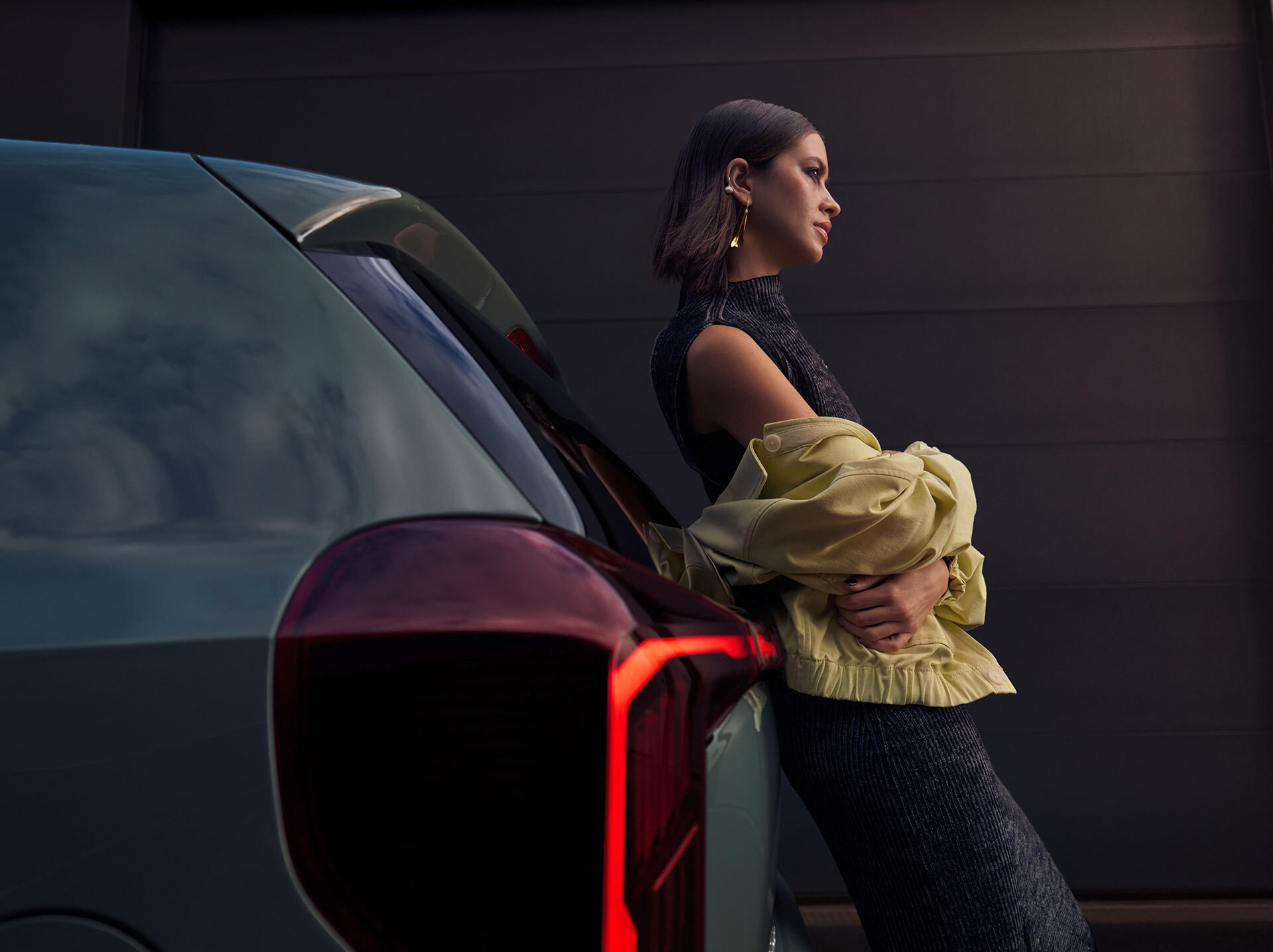 Kia Picanto in Metallic-Grün mit charakteristischer LED-Heckleuchten-Signatur, junge Frau in stilvollem schwarzen Kleid und gelber Jacke lehnt am Fahrzeug