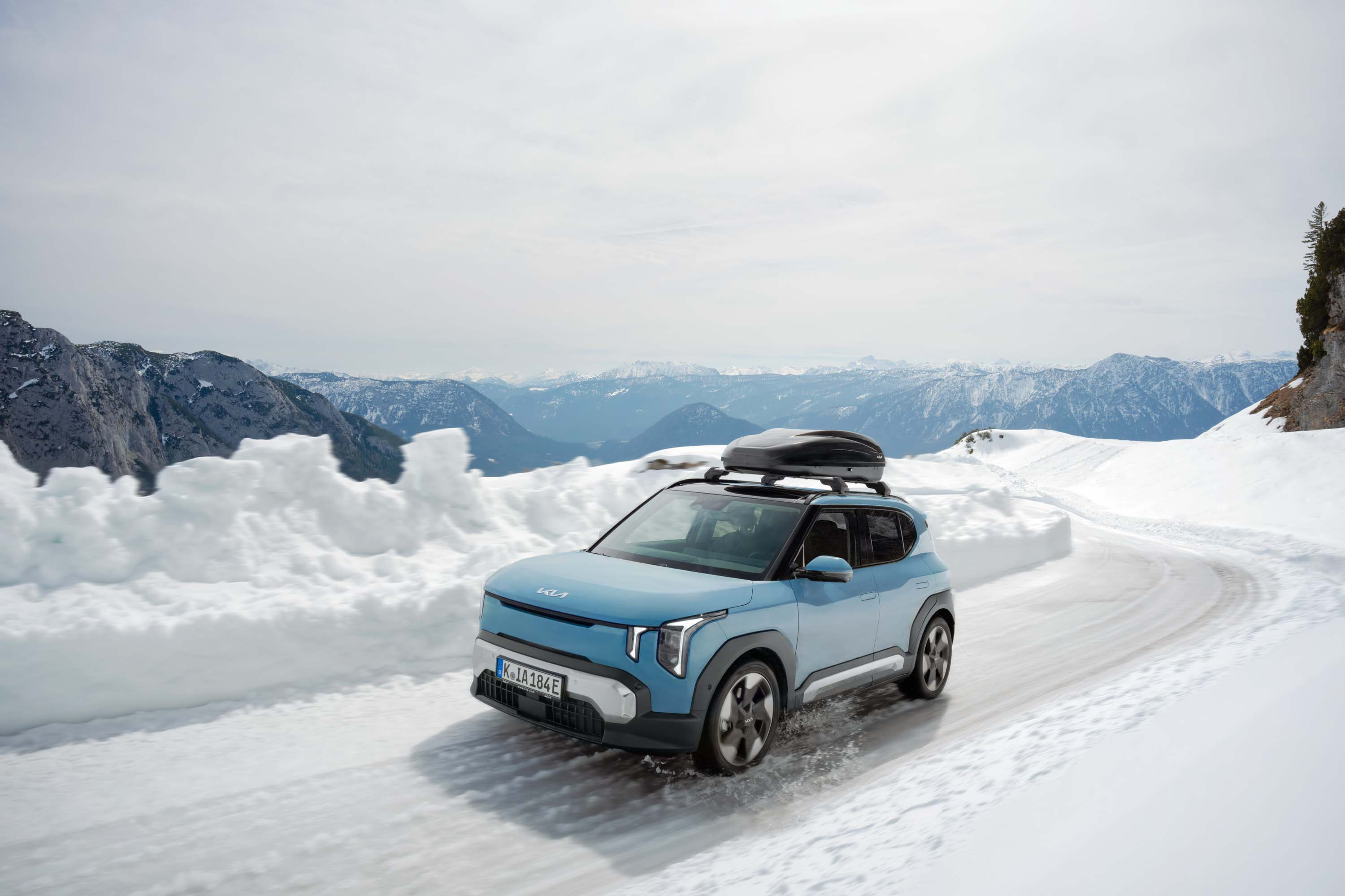 Kia EV2 in Hellblau mit Dachbox fährt auf einer verschneiten Bergstraße