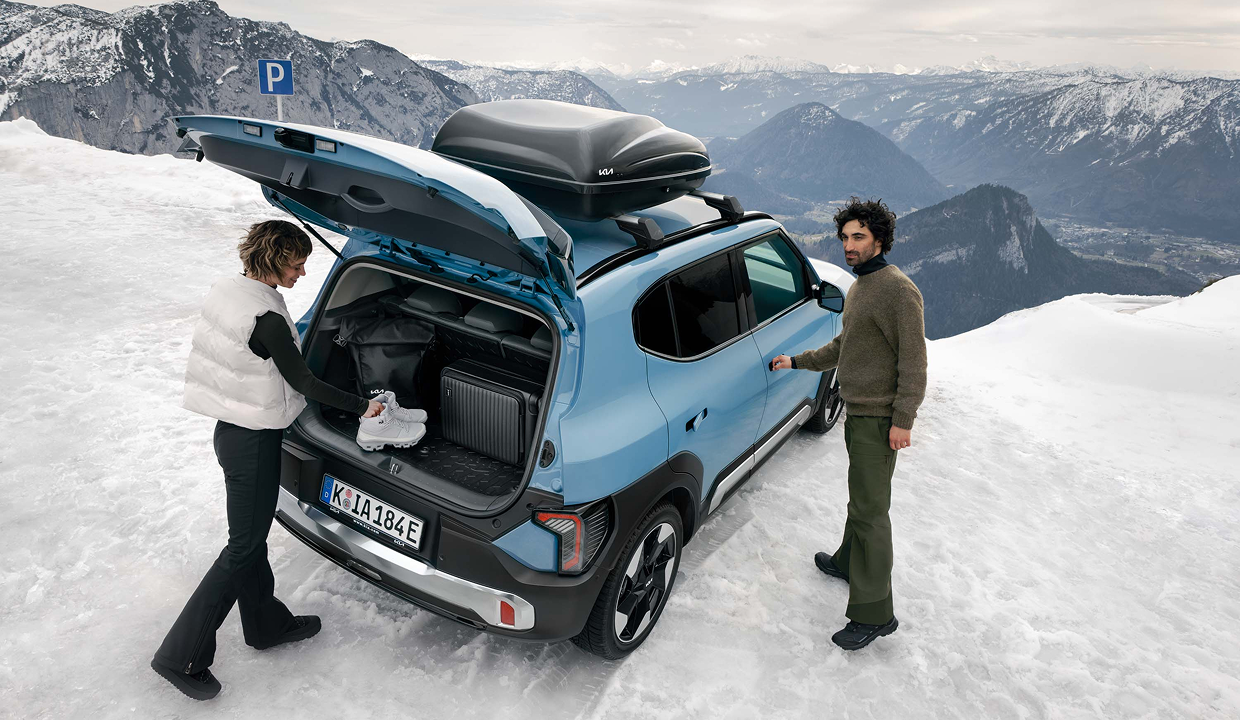 Zwei Personen beladen einen Kia EV2 mit Dachbox auf einem verschneiten Parkplatz in den Bergen